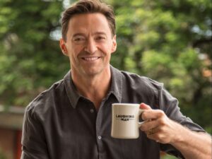 Hugh Jackman smiling and holding a cup of laughing man coffee. His own coffee brand.